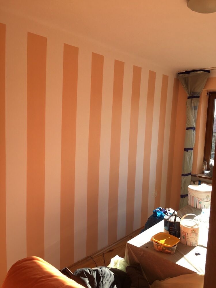 Pomarańczowo-białe pionowe pasy na ścianie pokoju, widoczne wiaderka z farbą i zabezpieczone okno folią malarską, w trakcie prac wykończeniowych.