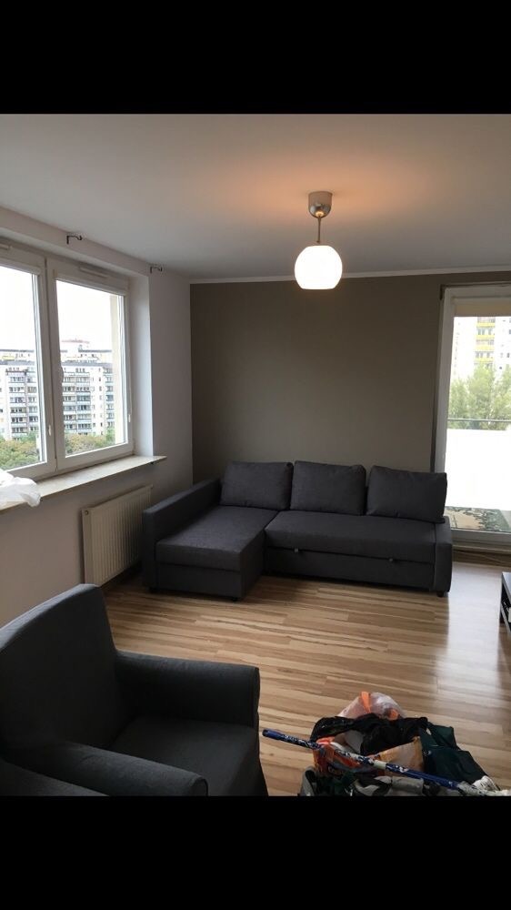 Szary narożnik z funkcją spania w pokoju z panelami podłogowymi i ścianą pomalowaną na szaro, widok na miasto z okna, w rogu sterta ubrań i kij hokejowy.