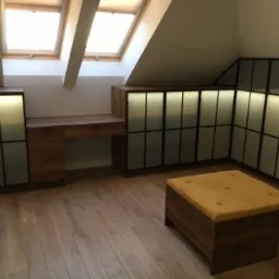 Pokój na poddaszu z zabudową: szafy z podświetleniem i szklanymi frontami, biurko z drewna oraz pufa z żółtym siedziskiem, drewniana podłoga, okna dachowe.