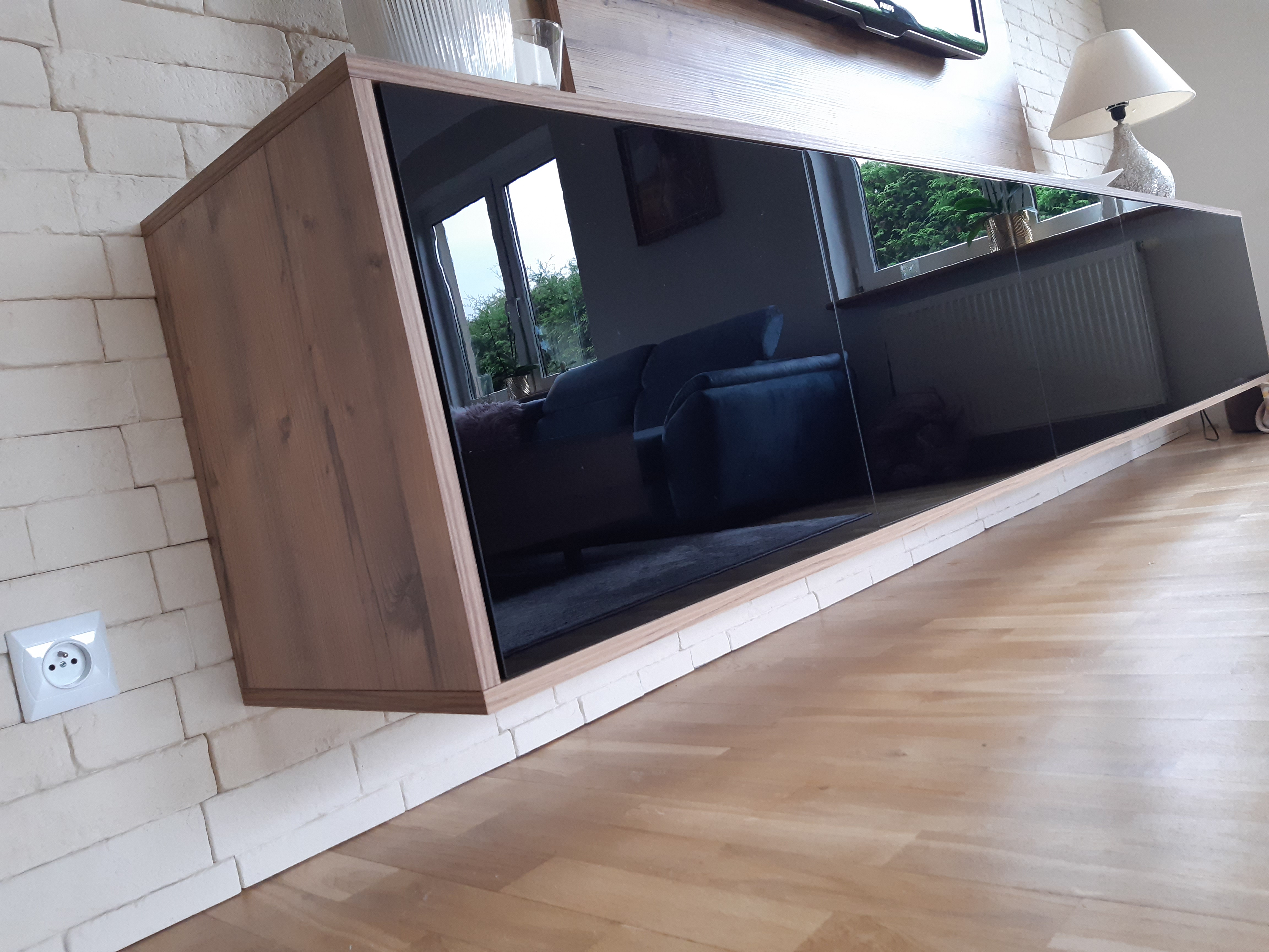 Wisząca szafka RTV z drewnianymi bokami i czarnymi, szklanymi frontami, zamontowana na ścianie z białej cegły dekoracyjnej, odbijająca wnętrze salonu. Na blacie lampa i dekoracje.