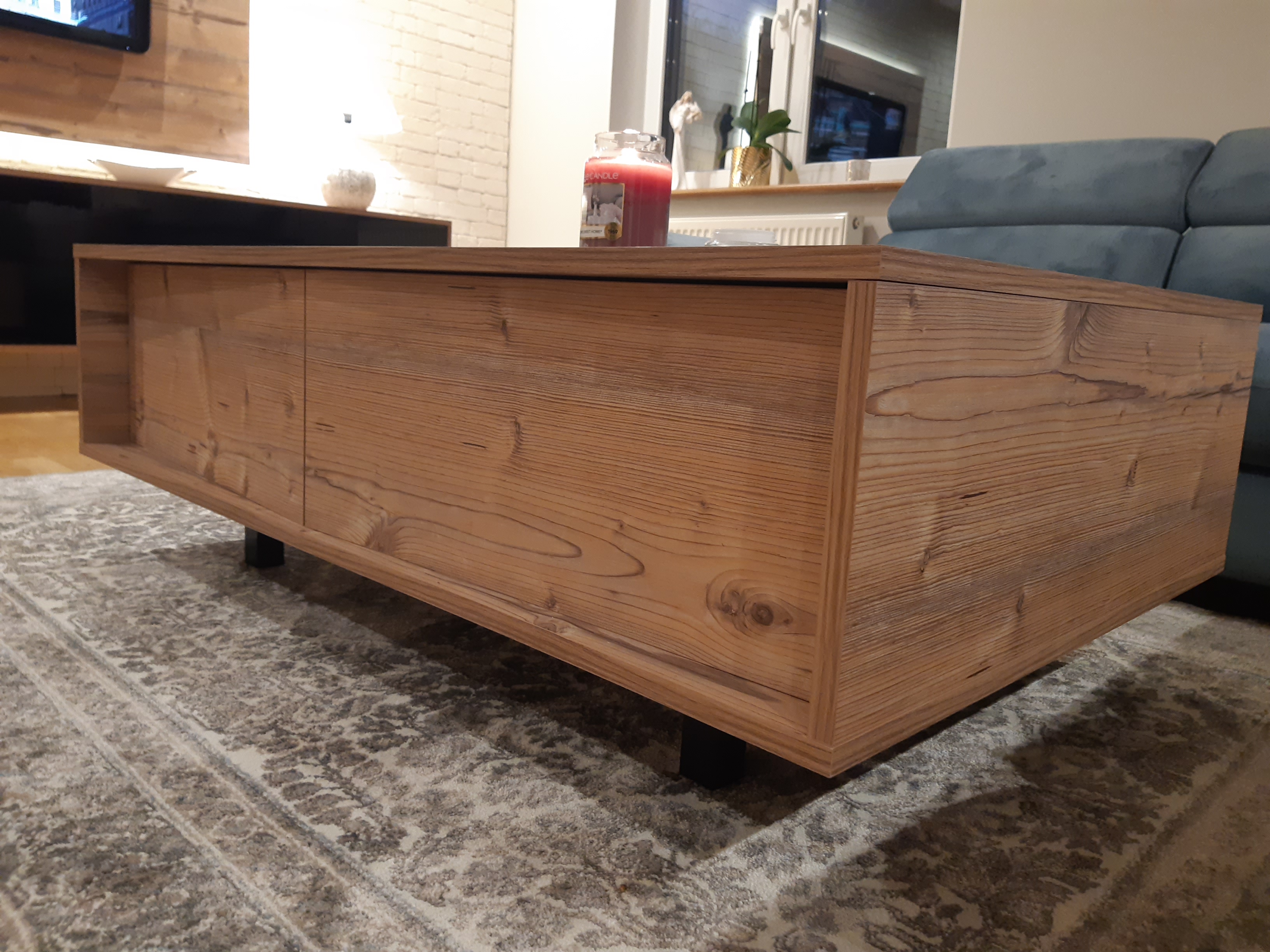 Drewniany stolik kawowy z szufladami, wykonany z jasnego drewna z widocznym usłojeniem, stoi na czarnych nóżkach na wzorzystym dywanie w salonie.