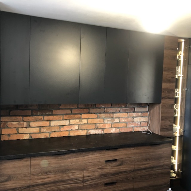 Nowoczesna kuchnia z czarnymi szafkami górnymi, blatem z ciemnego kamienia i dolnymi szafkami z forniru drewnopodobnego, podkreślona ścianą z cegły klinkierowej i oświetleniem LED w witrynie.