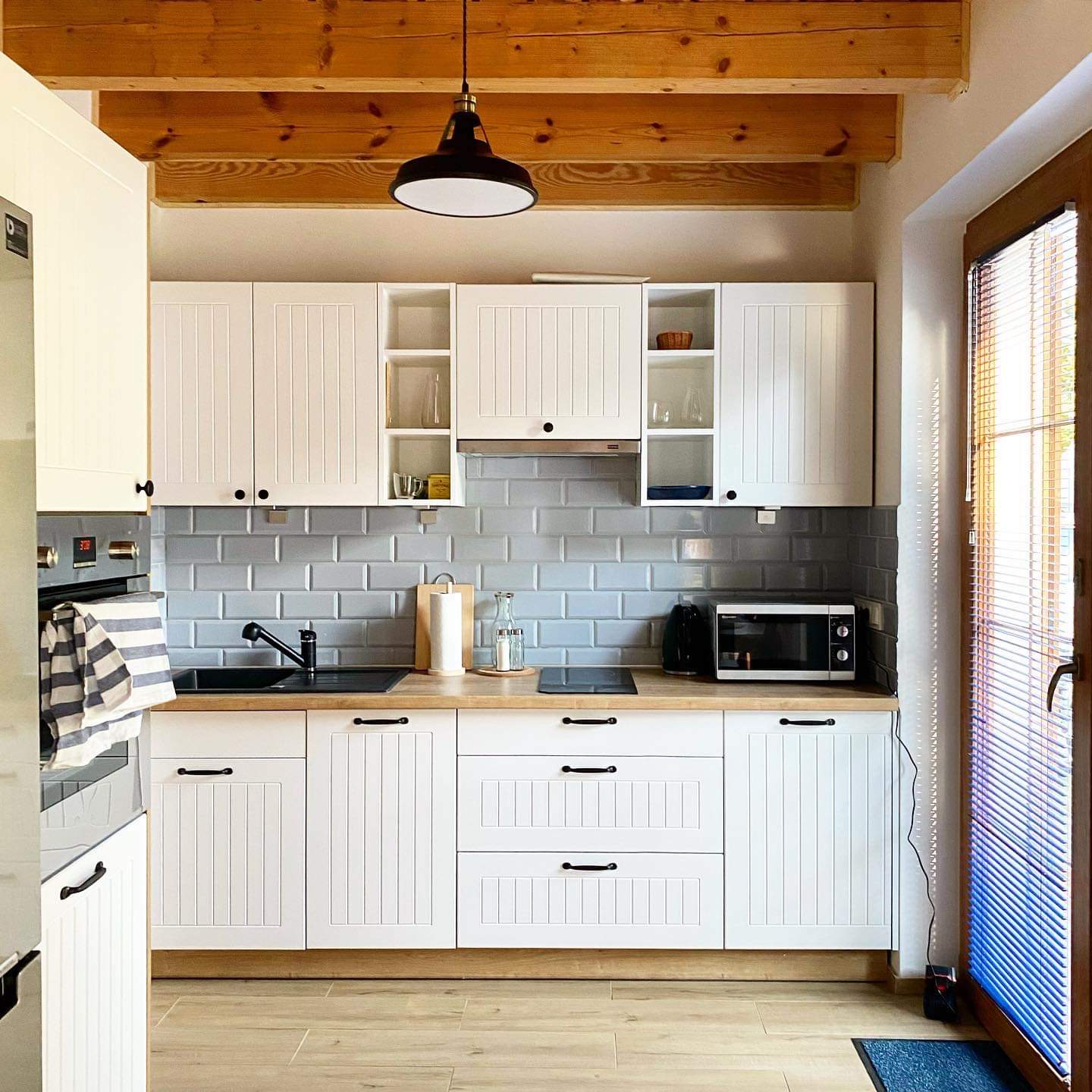 Jasna kuchnia z białymi szafkami z frezowanymi frontami, drewnianym blatem, szarymi płytkami metra i drewnianymi belkami stropowymi, czarnym zlewem i armaturą, oraz czarną lampą wiszącą.