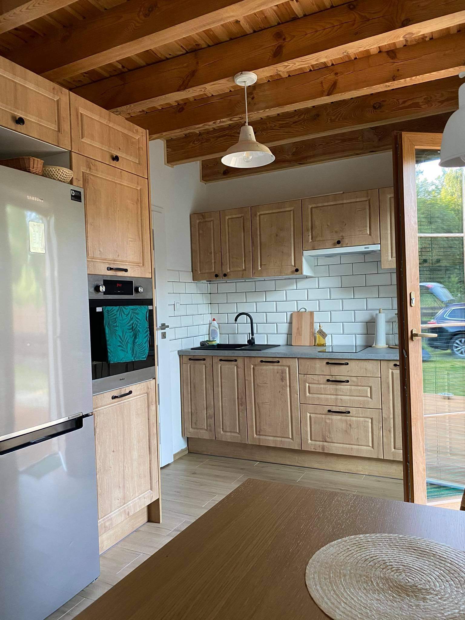 Jasna kuchnia z drewnianymi szafkami, czarnym zlewem i drewnianym stropem, widziana z perspektywy stołu z podkładką.