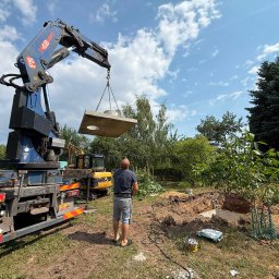 D&N SERWIS Dawid Nowik - Niebieski żuraw PM 48 sp na ciężarówce unosi betonową płytę z otworem nad wykopem, mężczyzna w szarym T-shircie nadzoruje prace, w tle koparka.