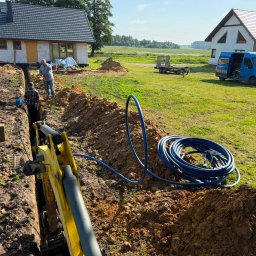 D&N SERWIS Dawid Nowik - Dwaj pracownicy układają niebieskie rury w świeżo wykopanym rowie, z widocznym ramieniem koparki i częściowo zbudowanym domem w tle, w słoneczny dzień.