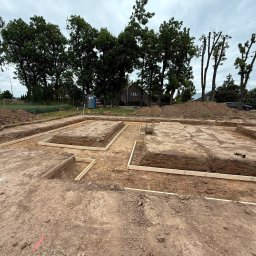 D&N SERWIS Dawid Nowik - Świeżo wykopane ławy fundamentowe pod budynek mieszkalny, z widocznym drewnianym szalowaniem i hałdami ziemi. W tle drzewa, przenośna toaleta i dom.