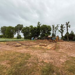 D&N SERWIS Dawid Nowik - Wykopane fundamenty pod budynek z drewnianym szalunkiem na błotnistym placu budowy, w tle żółta koparka i przycięte drzewa pod zachmurzonym niebem.