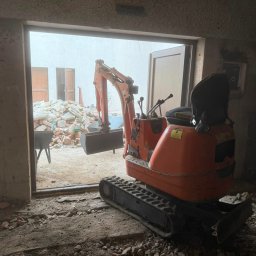 D&N SERWIS Dawid Nowik - Pomarańczowa mini-koparka gąsienicowa w ciemnym wnętrzu budynku, z widocznym przez duży otwór wyjściowy stosem gruzu budowlanego na jasnym podwórku.