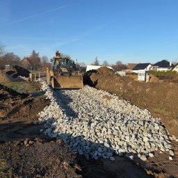 D&N SERWIS Dawid Nowik - Żółta koparko-ładowarka Caterpillar rozkłada jasne, nieregularne kruszywo na przygotowanym podłożu, otoczonym hałdami ziemi. W tle domy i błękitne niebo.