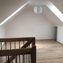 Poddasze po remoncie z drewnianą podłogą, białymi ścianami, oknem dachowym i schodami z drewnianą balustradą z czarnymi szczebelkami, widok z góry schodów.