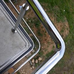 Tomasz Wasilewski - Błyszcząca, metalowa konstrukcja zabezpieczająca balkon, z widocznym odbiciem nieba na polerowanej powierzchni, otoczona zielenią i elementami placu budowy.