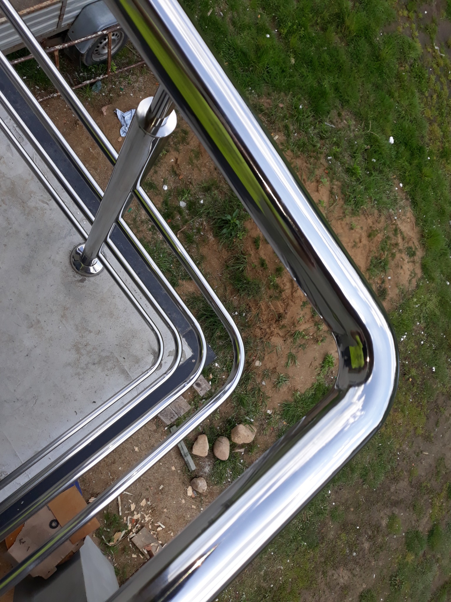 Błyszcząca, metalowa konstrukcja zabezpieczająca balkon, z widocznym odbiciem nieba na polerowanej powierzchni, otoczona zielenią i elementami placu budowy.