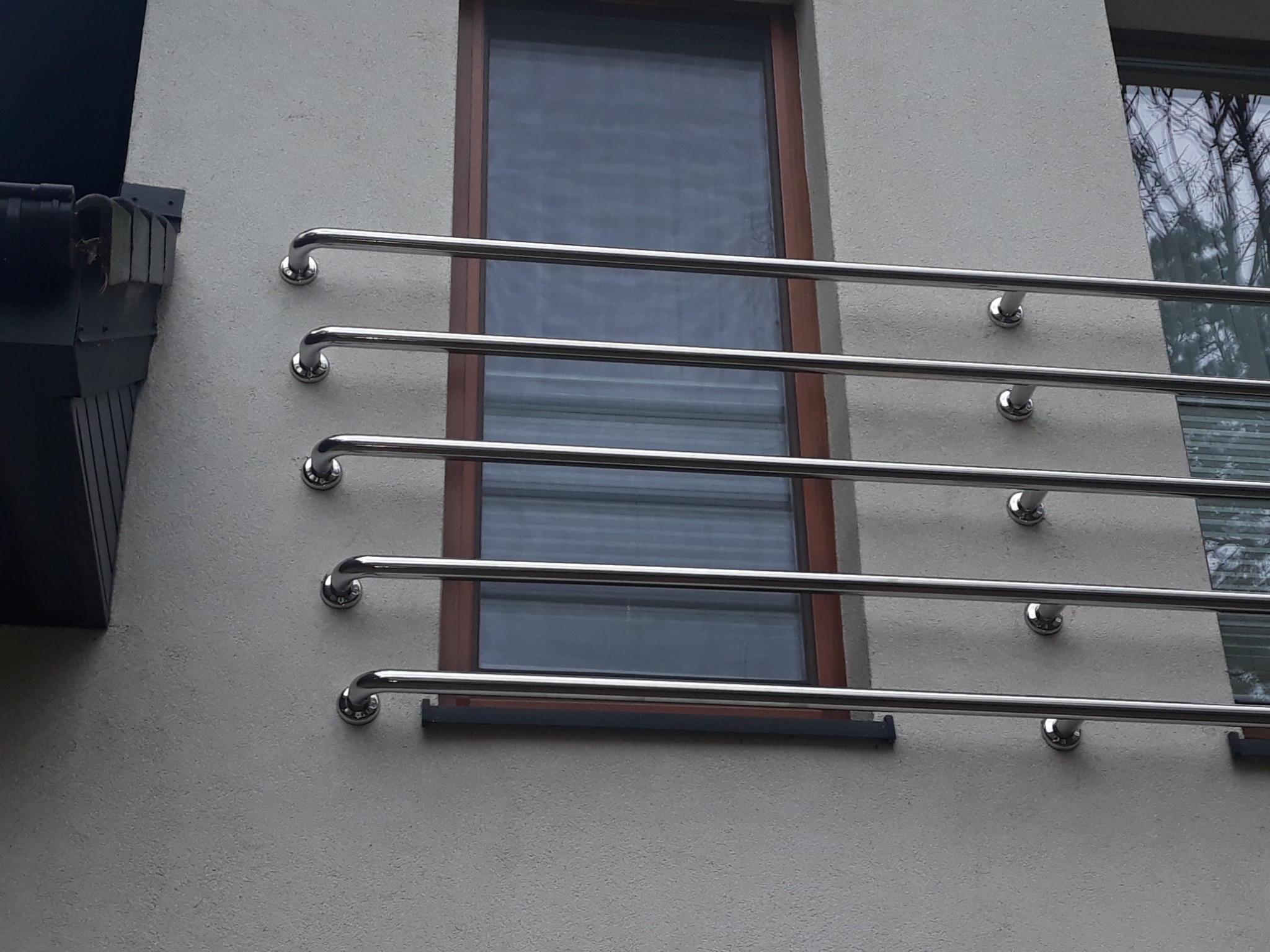 Zewnętrzna, metalowa konstrukcja ochronna z polerowanych rur, zamocowana do elewacji budynku, widoczna obok okna z drewnianą ramą.