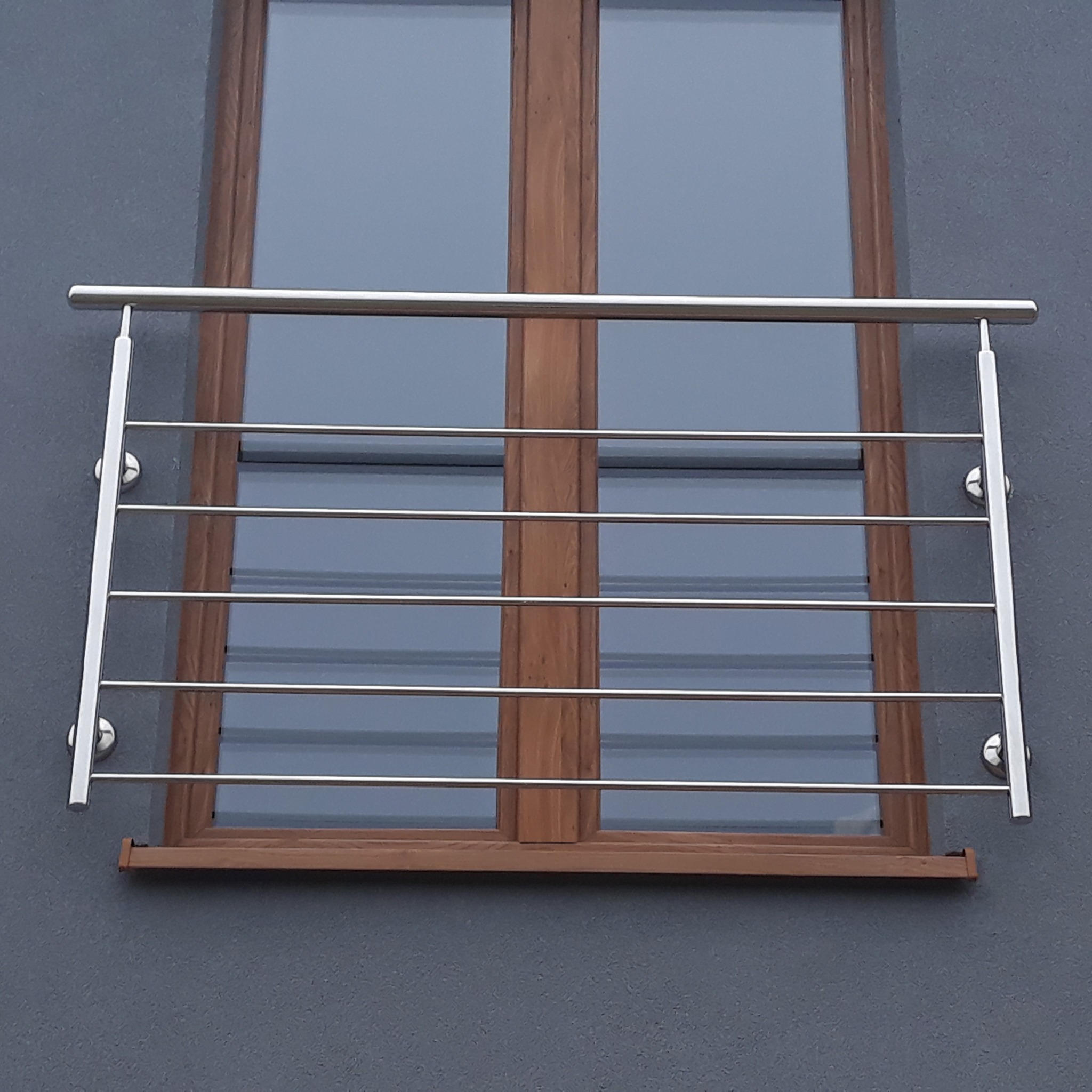 Nierdzewna barierka okienna z poziomymi prętami, zamocowana do szarej elewacji pod drewnianym oknem.