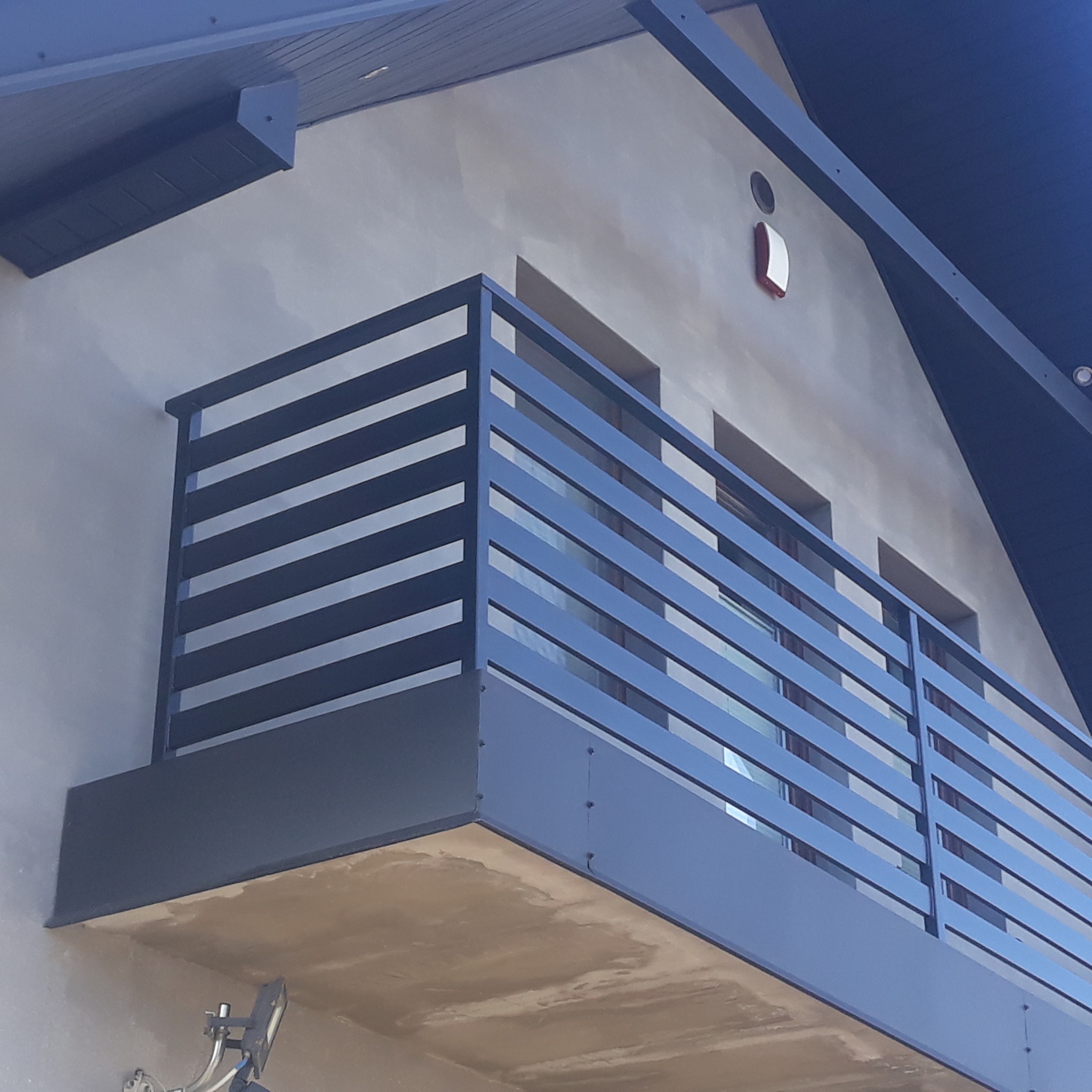 Balkon z nowoczesną, poziomą konstrukcją zabezpieczającą w kolorze grafitowym, zamontowaną na tle szarej elewacji budynku z widocznym fragmentem dachu i anteną satelitarną.