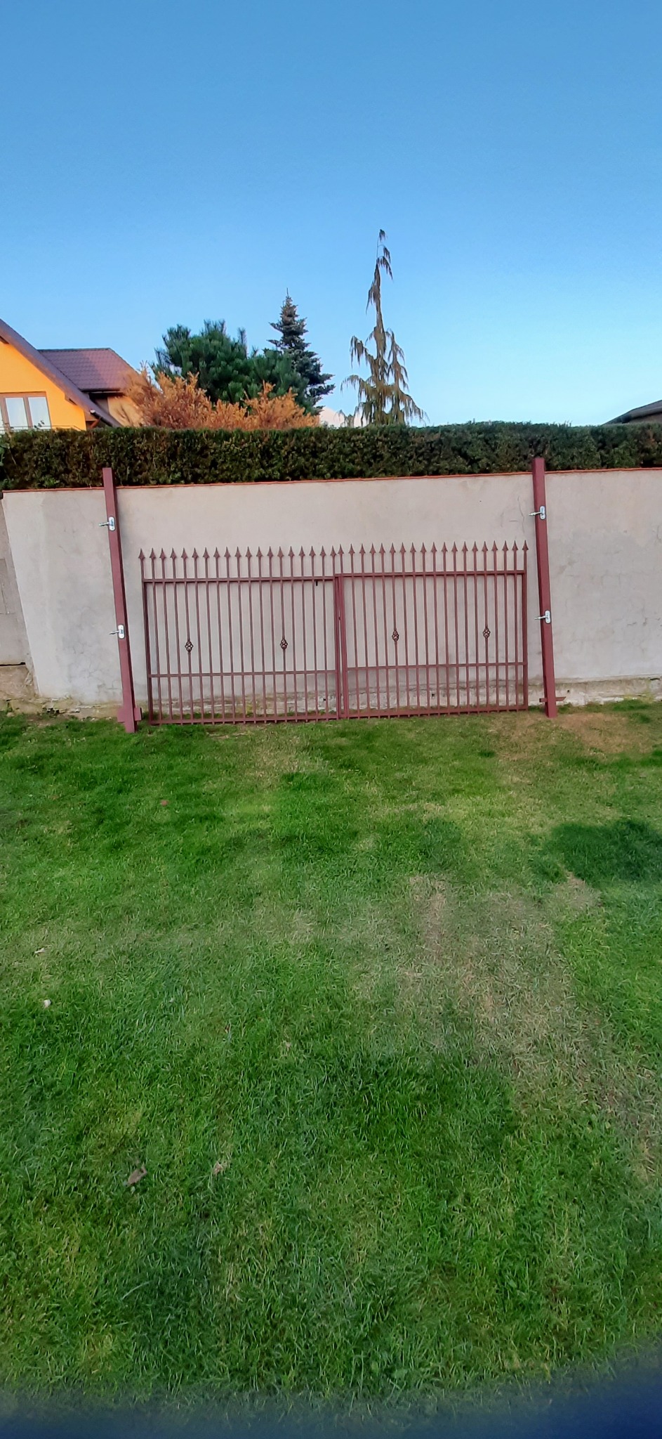 Dwie części metalowej bramy wjazdowej, o ciemnobrązowym kolorze, oparte o słupki i ścianę posesji, na tle zielonego trawnika i żywopłotu.