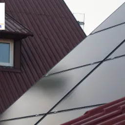 Częściowy widok paneli słonecznych na dachu domu z brązową dachówką i oknem mansardowym, logo firmy w lewym górnym rogu.