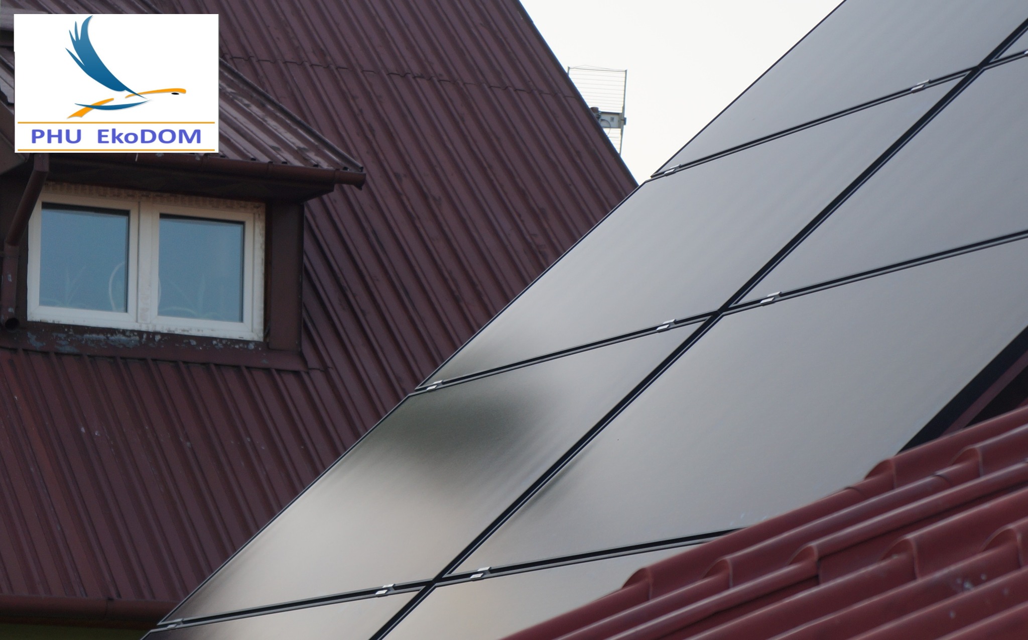 Częściowy widok paneli słonecznych na dachu domu z brązową dachówką i oknem mansardowym, logo firmy w lewym górnym rogu.