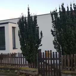 Nowoczesny dom jednorodzinny z drewnianą elewacją, białymi ścianami i dużymi oknami w czarnych ramach, widziany zza drewnianego płotu.