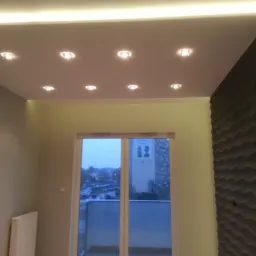 Pokój z podwieszanym sufitem z oświetleniem punktowym i listwą LED, ściana z dekoracyjną strukturą 3D, widok na balkon i blok mieszkalny za oknem.