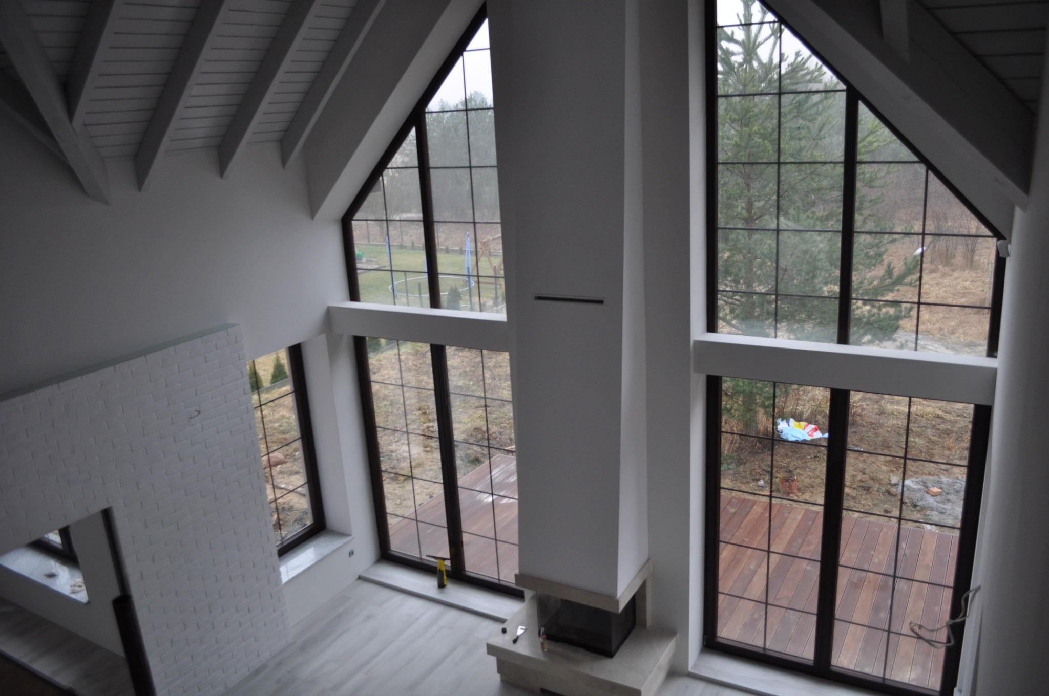 Wysokie wnętrze domu z drewnianą podłogą widoczną przez duże okna, białe ściany, drewniany sufit z belkami i kominek z jasnego kamienia.