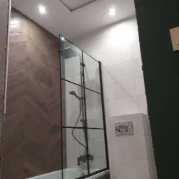 Nowoczesna łazienka z kabiną prysznicową typu walk-in z czarnymi profilami, drewnopodobnymi płytkami ułożonymi w jodełkę na ścianie oraz białymi płytkami na pozostałych ścianach, podwieszany sufit...