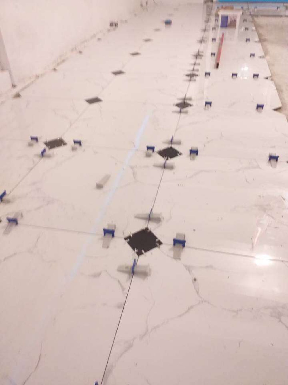 Układanie białych, marmurkowych płytek podłogowych z użyciem czarnych znaczników i niebieskich klipsów poziomujących. Widoczna linia laserowa wyznaczająca kierunek układania.