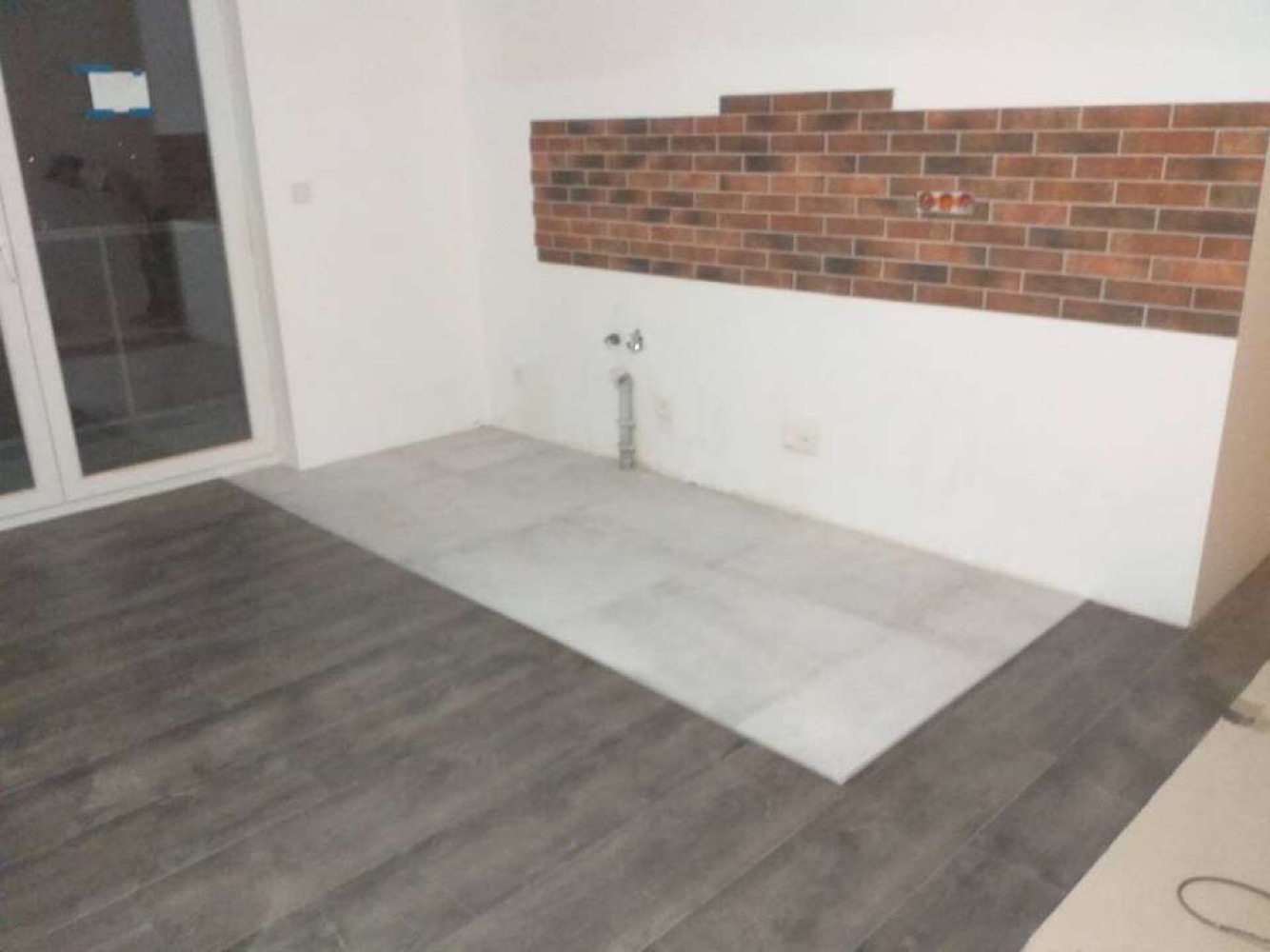 Podłoga w trakcie układania płytek: część z szarymi panelami imitującymi drewno, część z jasnymi płytkami; na ścianie fragment dekoracyjnej cegły i wystające rury.