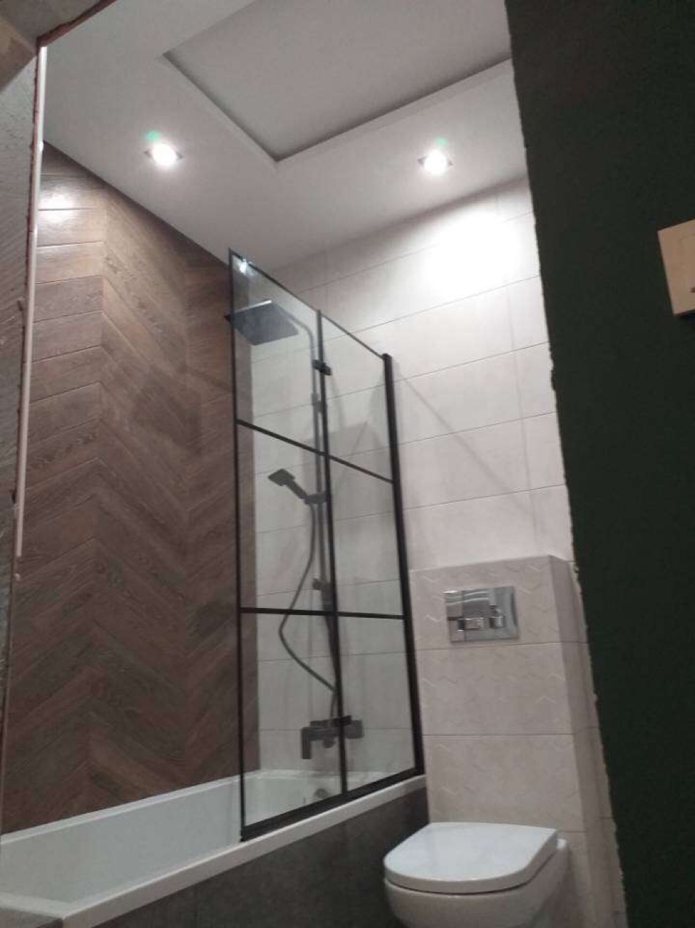 Nowoczesna łazienka z kabiną prysznicową typu walk-in z czarnymi profilami, drewnopodobnymi płytkami ułożonymi w jodełkę na ścianie oraz białymi płytkami na pozostałych ścianach, podwieszany sufit...