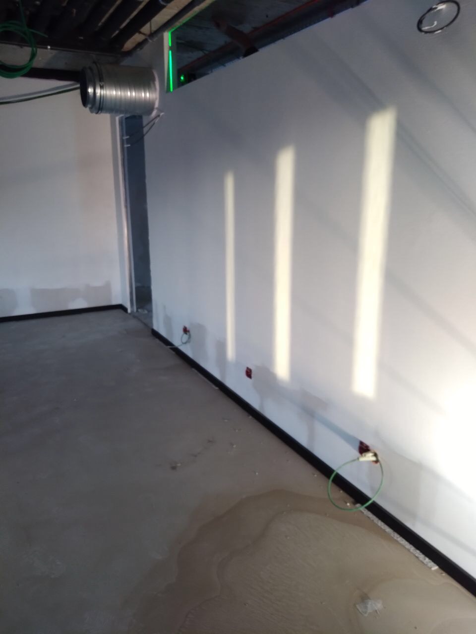 Wnętrze w trakcie prac wykończeniowych, widoczne szpachlowane ściany z widocznymi śladami po łatach, surowa podłoga z plamami, wystające kable elektryczne, oraz metalowa rura wentylacyjna pod sufitem.