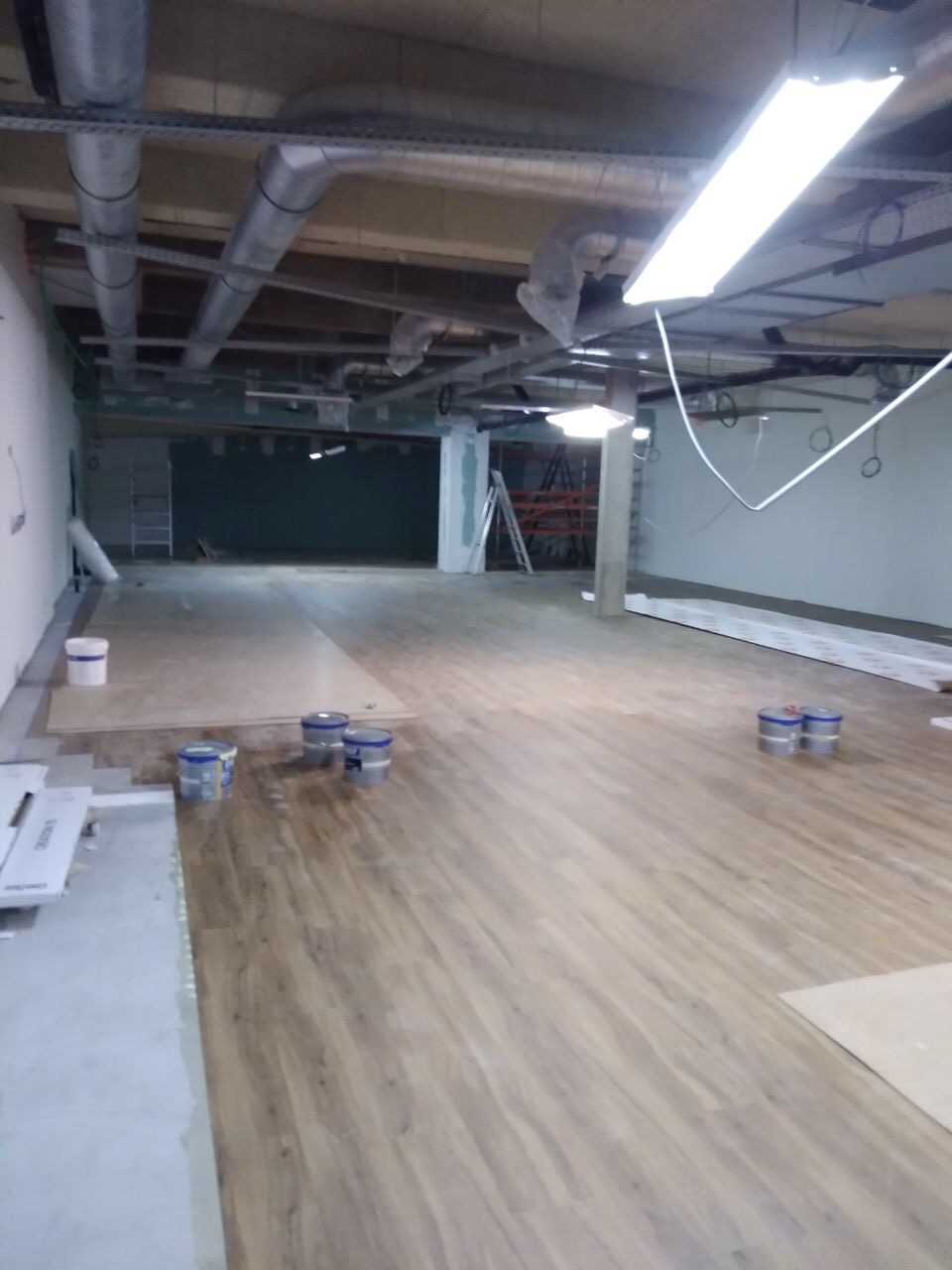 Układanie paneli podłogowych w dużym, surowym pomieszczeniu z widocznymi instalacjami pod sufitem i puszkami farby na podłodze.