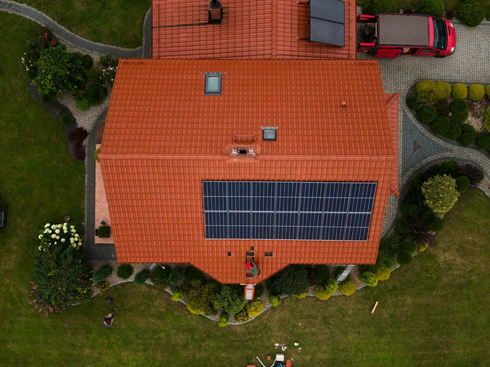 Widok z lotu ptaka na dom z czerwoną dachówką, na której zamontowane są panele słoneczne; pracownik na dachu przy panelach, czerwony samochód dostawczy zaparkowany obok, zadbany ogród.