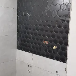 Ściana w łazience z częściowo ułożonymi czarnymi heksagonalnymi płytkami z delikatną fakturą, poniżej szare płytki wielkoformatowe, widoczne przygotowane punkty instalacyjne.