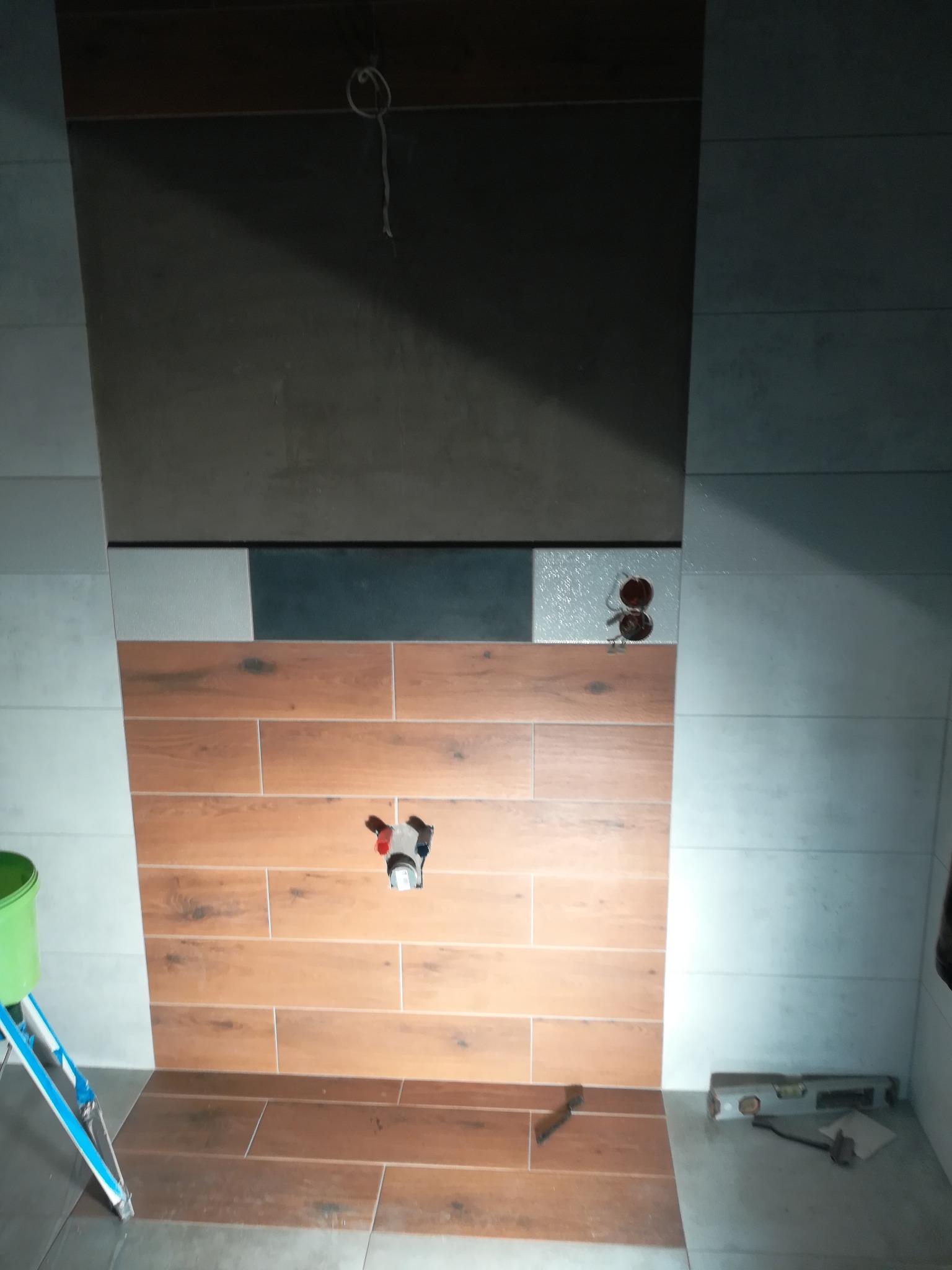 Montaż płytek imitujących drewno w kabinie prysznicowej, widoczne narzędzia, poziomica, drabinka i wiadro, surowy beton na górze ściany.