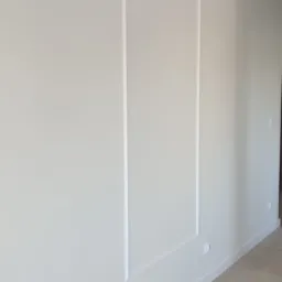 Ściana w jasnym odcieniu z dekoracyjnymi, pionowymi wgłębieniami tworzącymi prostokątne panele. Minimalistyczny design, widoczne subtelne załamania światła na powierzchni.