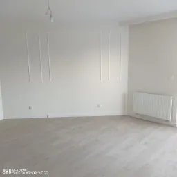 Jasne, puste pomieszczenie po remoncie z panelami podłogowymi w odcieniach szarości i białymi ścianami z ozdobnymi listwami, widoczny grzejnik.