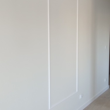 Ściana w jasnym odcieniu z dekoracyjnymi, pionowymi wgłębieniami tworzącymi prostokątne panele. Minimalistyczny design, widoczne subtelne załamania światła na powierzchni.