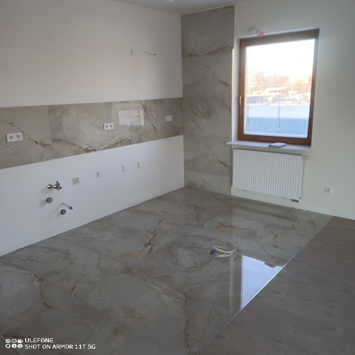 Wnętrze pokoju po remoncie: położona podłoga i częściowo płytki ścienne imitujące marmur, widoczne przygotowane instalacje elektryczne i hydrauliczne, grzejnik pod oknem.