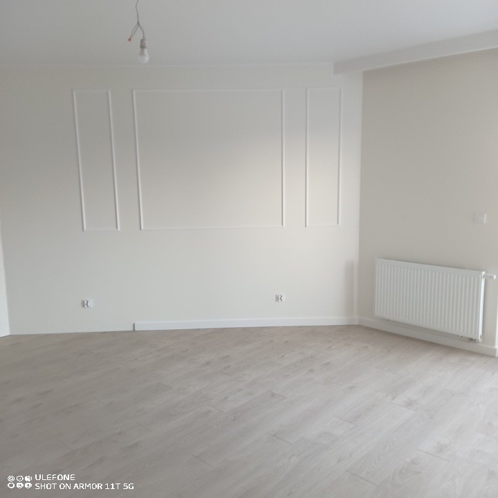 Jasne, puste pomieszczenie po remoncie z panelami podłogowymi w odcieniach szarości i białymi ścianami z ozdobnymi listwami, widoczny grzejnik.