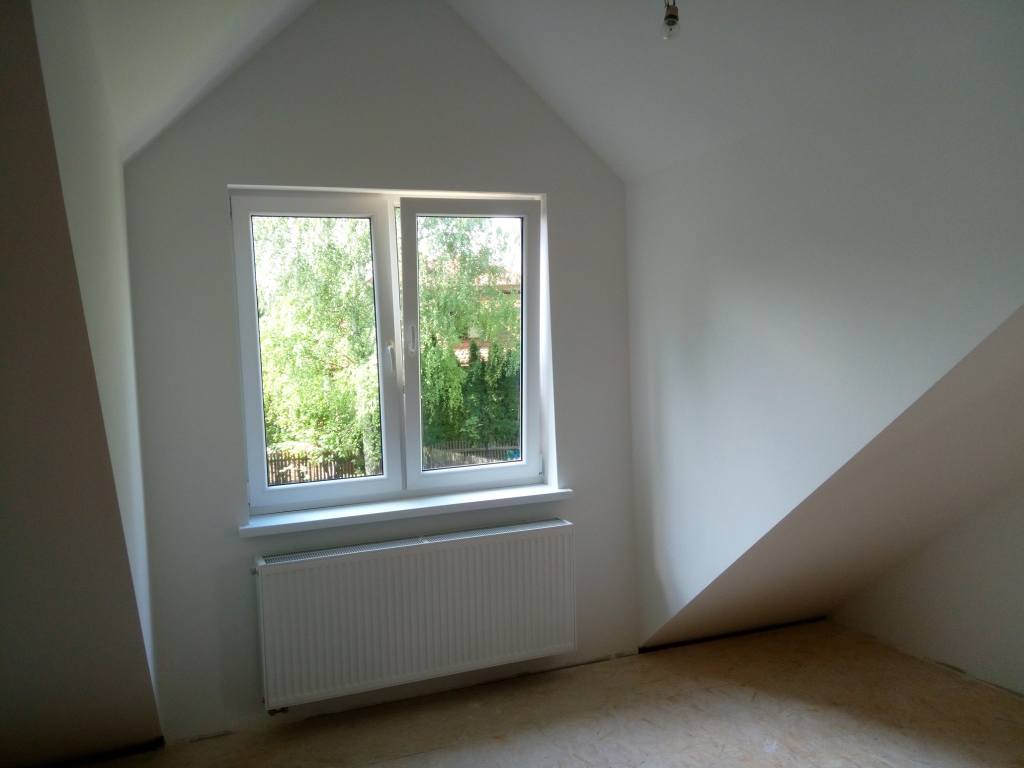 Poddasze po remoncie: białe ściany, okno z widokiem na zieleń, grzejnik pod oknem, surowa podłoga przed ułożeniem paneli.