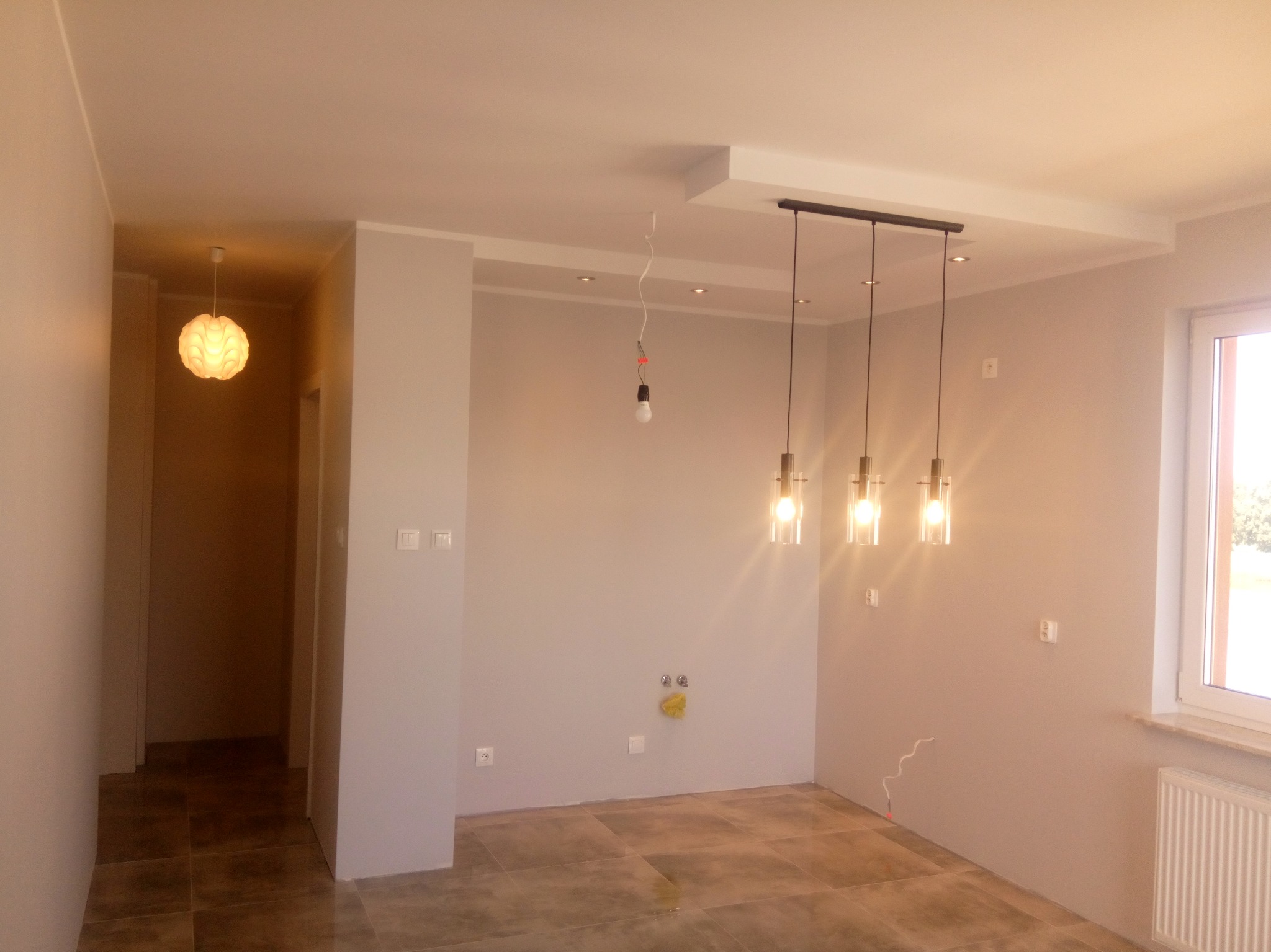 Wnętrze pokoju z wykończonymi ścianami w odcieniach szarości, z zamontowanymi trzema lampami wiszącymi na czarnej listwie, widoczne gniazdka elektryczne i wyprowadzone kable, podłoga wyłożona...