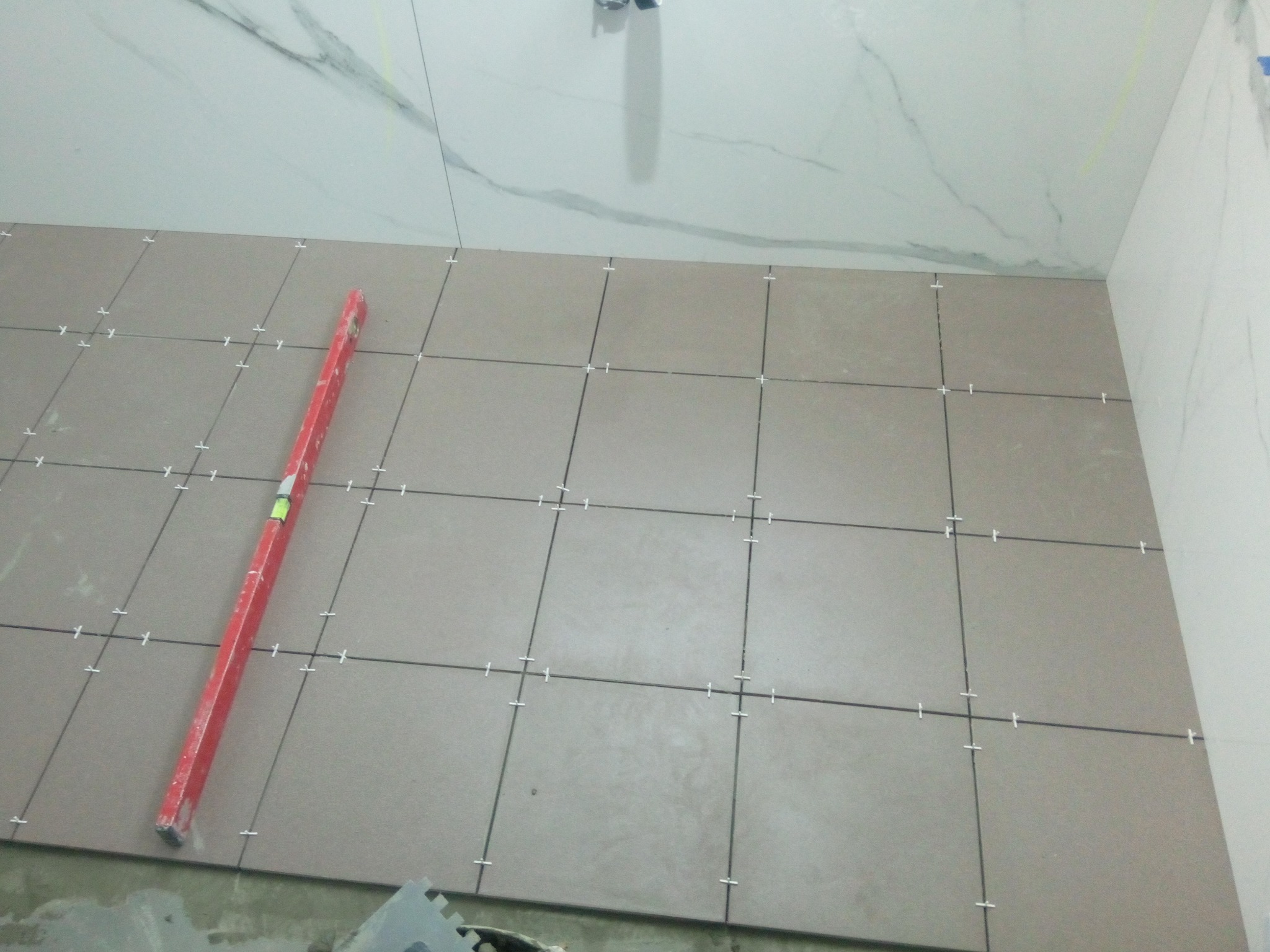 Ułożone płytki podłogowe z krzyżykami dystansowymi, poziomica przy ścianie, białe płytki ścienne imitujące marmur w tle.