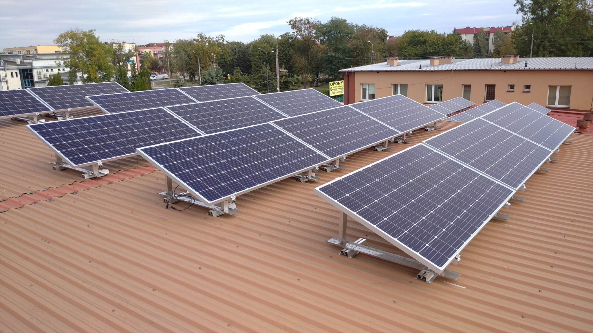 Rząd paneli słonecznych na metalowym dachu budynku, widok z góry, z widocznymi elementami montażowymi i otoczeniem miejskim w tle.