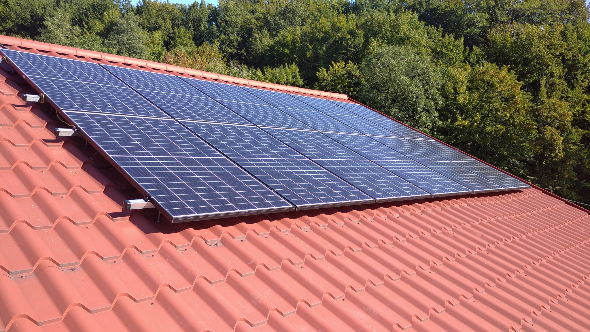 Panele słoneczne zamontowane na czerwonym dachu z dachówki ceramicznej, widok z dołu, w tle drzewa.