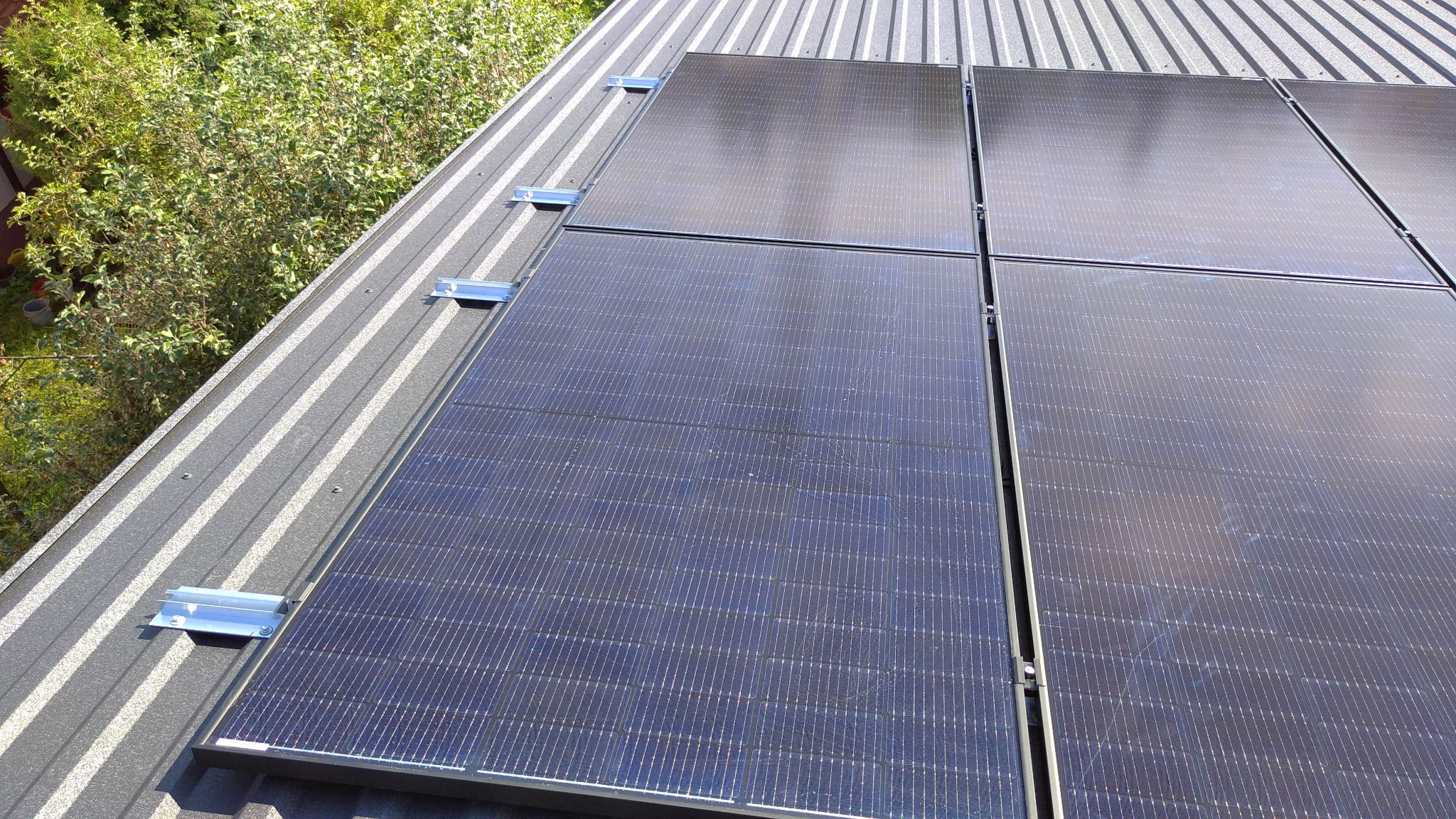 Panele słoneczne zamontowane na blaszanym dachu z widocznymi mocowaniami, w tle zieleń drzew.