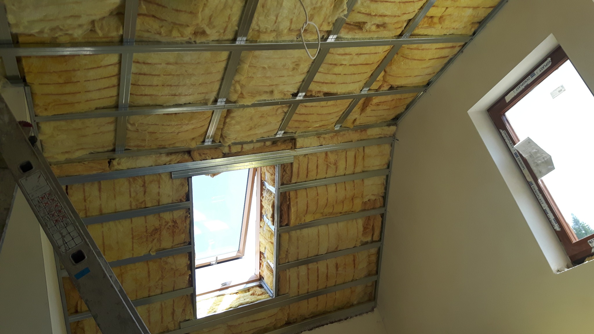 Montaż izolacji z wełny mineralnej w poddaszu z oknem dachowym, widoczne metalowe profile i częściowo otynkowane ściany, drabina przystawiona z lewej strony.