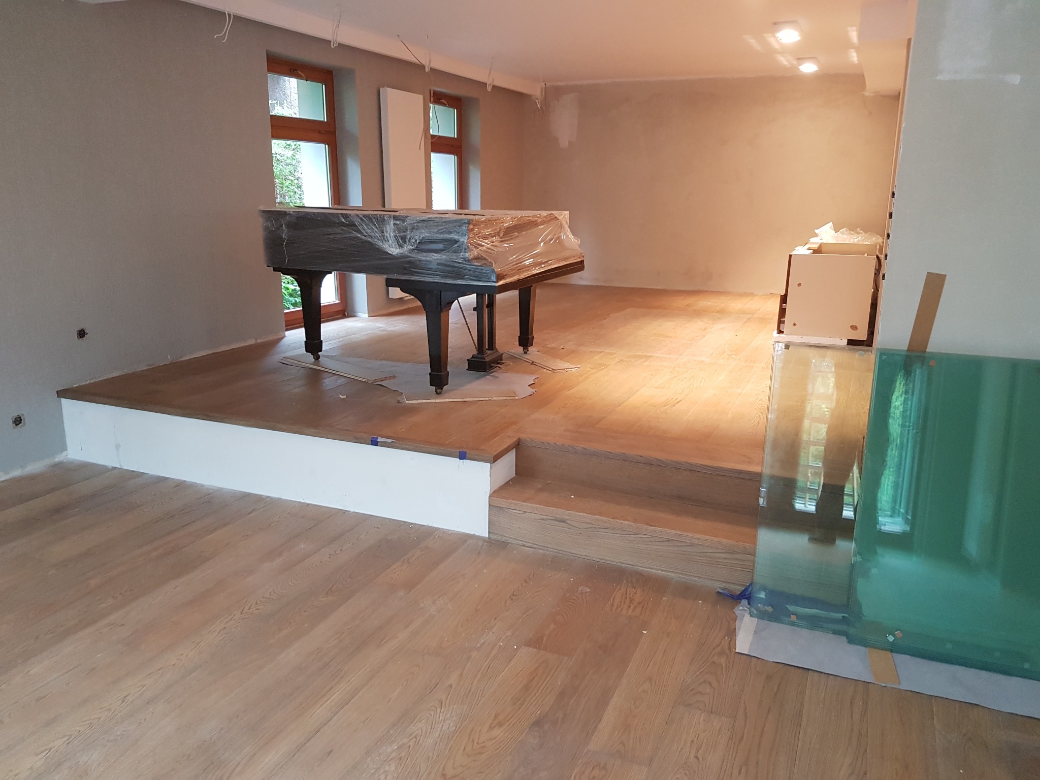Wnętrze w trakcie prac, z drewnianą podłogą, schodami i podestem; fortepian zabezpieczony folią, ściany przygotowane do malowania, widoczne okna i elementy instalacji elektrycznej.