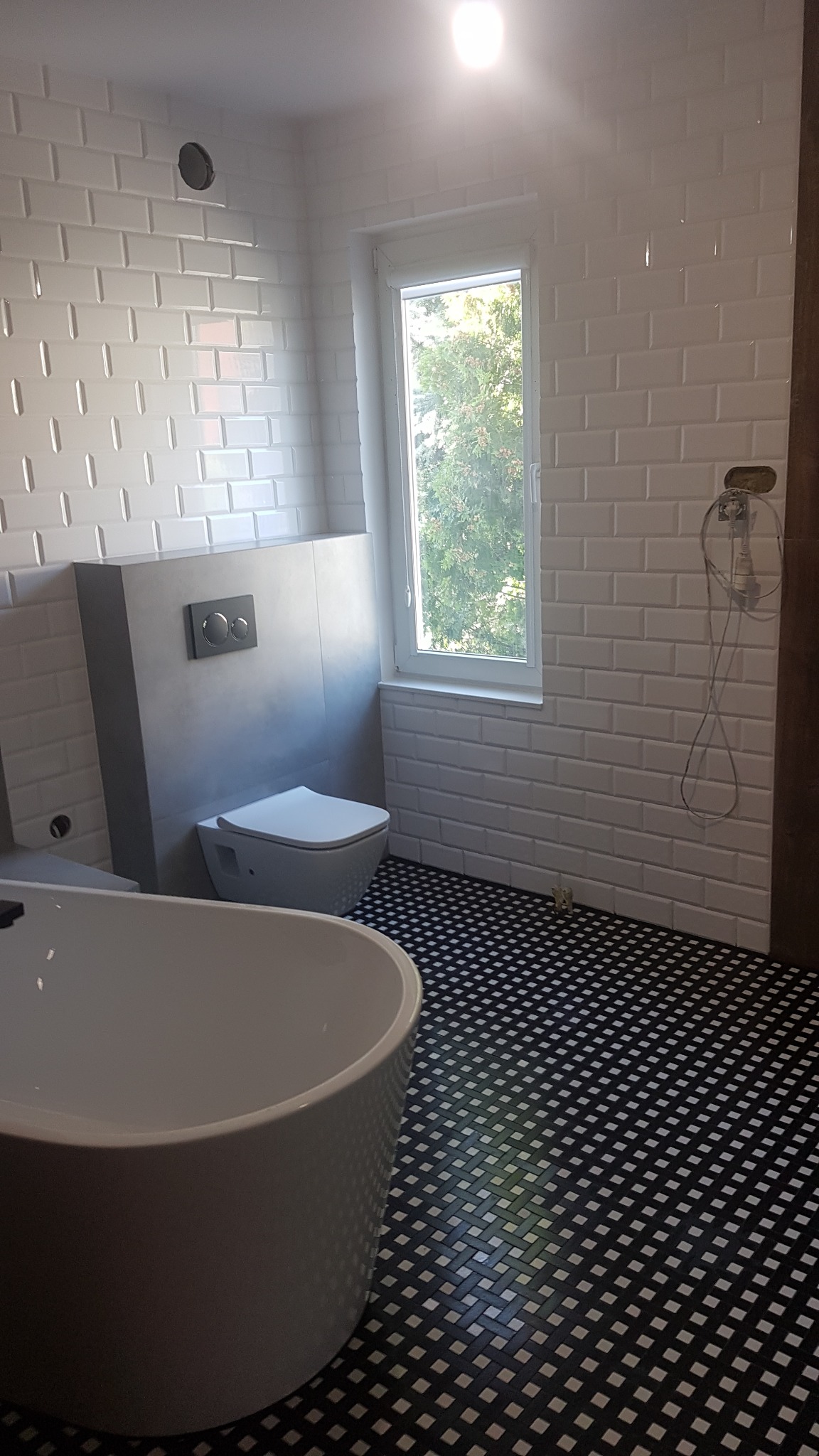 Nowoczesna łazienka z białą armaturą, białą cegiełkową glazurą i czarno-białą mozaiką na podłodze. Widoczna wanna wolnostojąca, toaleta podwieszana z szarym stelażem i okno z widokiem na zieleń.