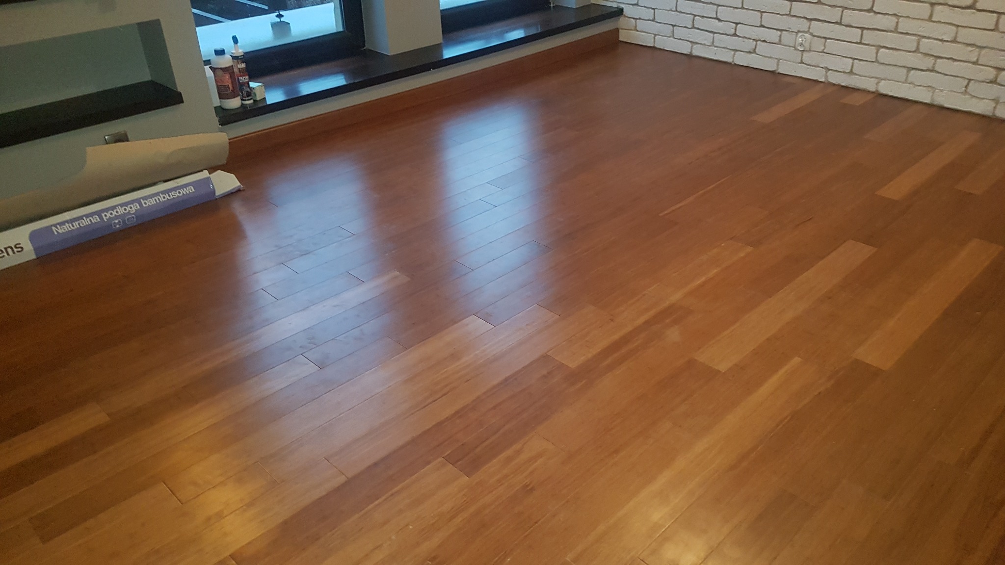 Ułożona podłoga bambusowa z widocznym opakowaniem paneli, biała ściana z cegieł w tle, okno z ciemną ramą.